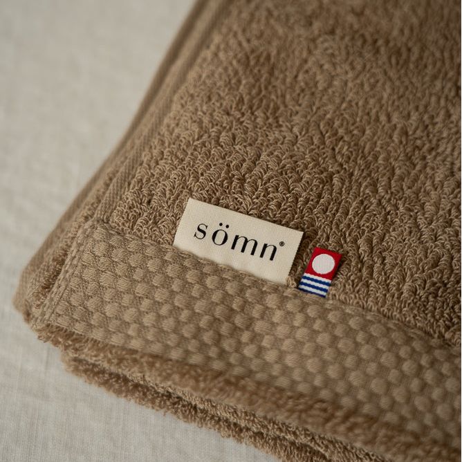 Serviette de bain en coton brun, bord texturé et étiquette sömn