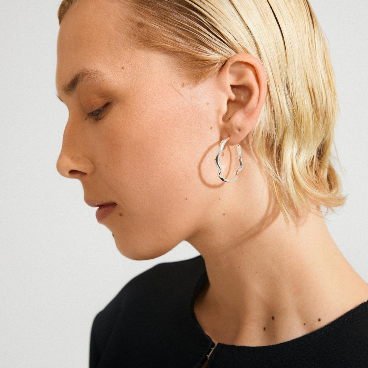 Boucles d’oreilles ondulées plaquées or, créoles ouvertes, portées avec cheveux relevés.