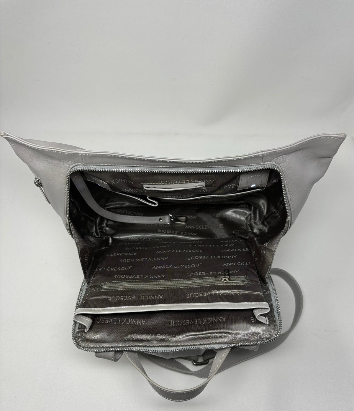 Sac à dos en cuir argent, intérieur doublé noir avec poches et sangles visibles.